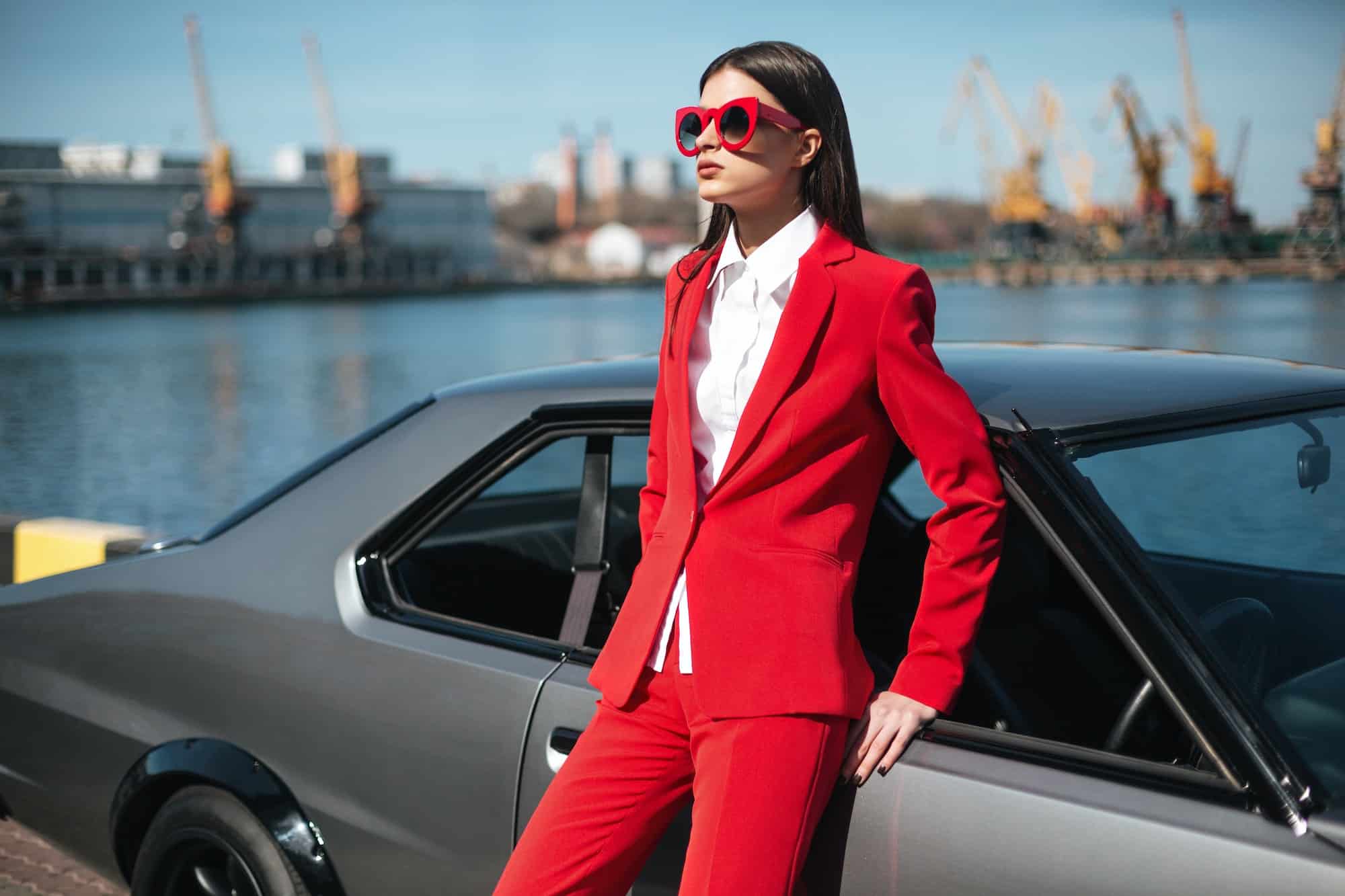 Fashion girl next to a retro sport car. Woman in a red suit and sunglasses waiting near classic car