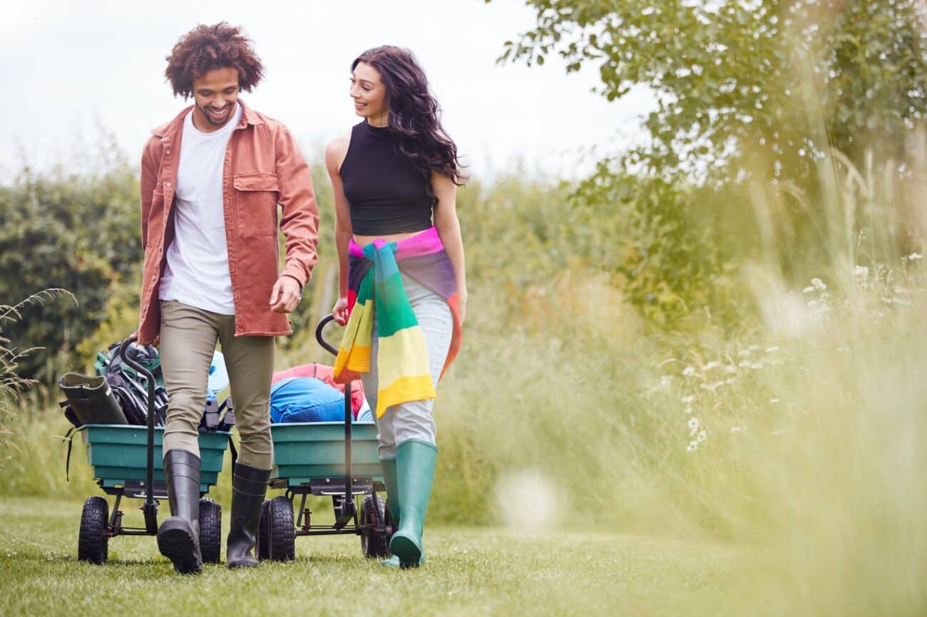 Young Couple Pulling Trolley Filled With Camping Equipment Through Field To Music Festival
